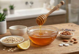 Ahşap bir masa üzerinde cam kaseden süzülen bal, yanında yulaf ezmesi, yarım limon, badem ve aspirinin bulunduğu, arka planda aydınlık bir banyonun göründüğü doğal cilt bakımı görseli.